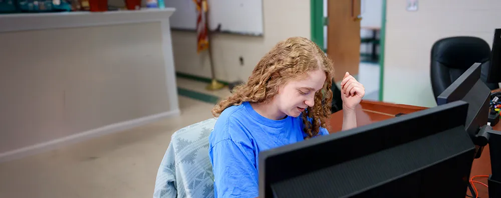 Student seated at a desk using dual computer monitors in a classroom with windows, posters, and computer equipment.