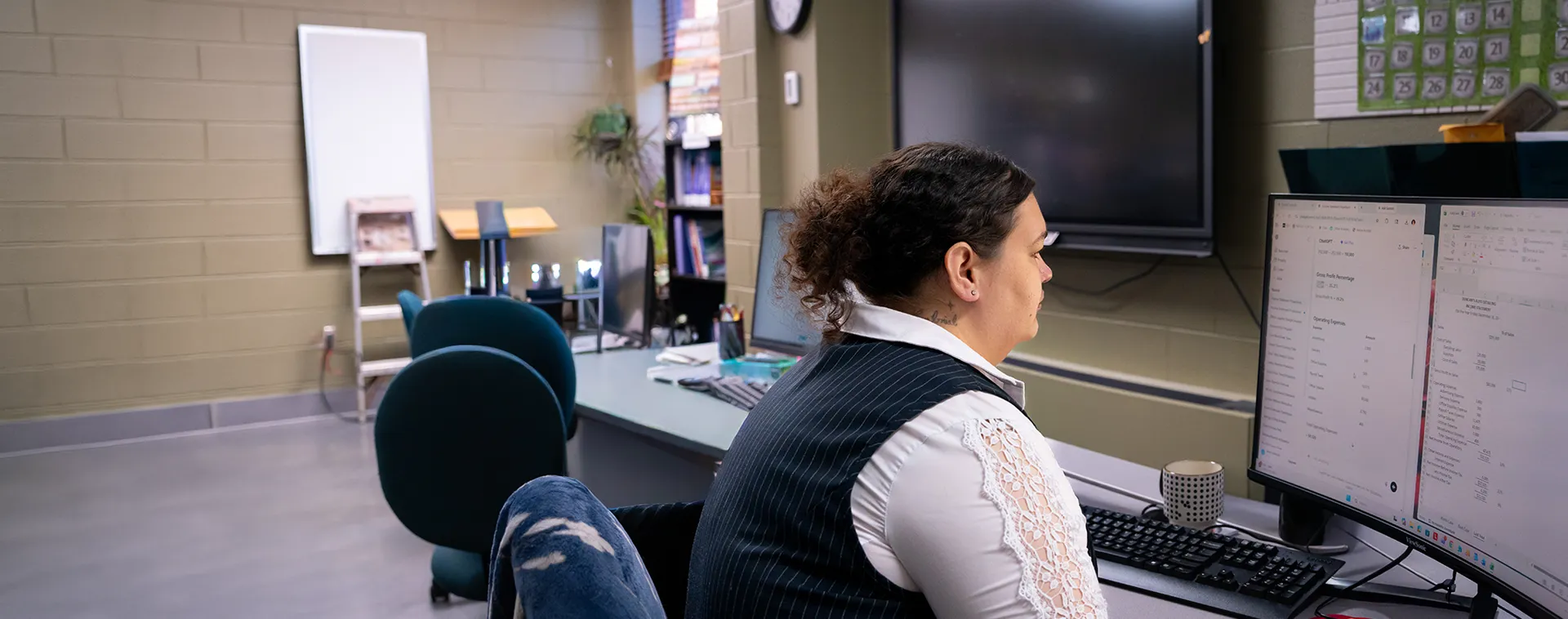 Student seated at a desk in a classroom, facing dual computer monitors with chairs, shelves, and a whiteboard in the background
