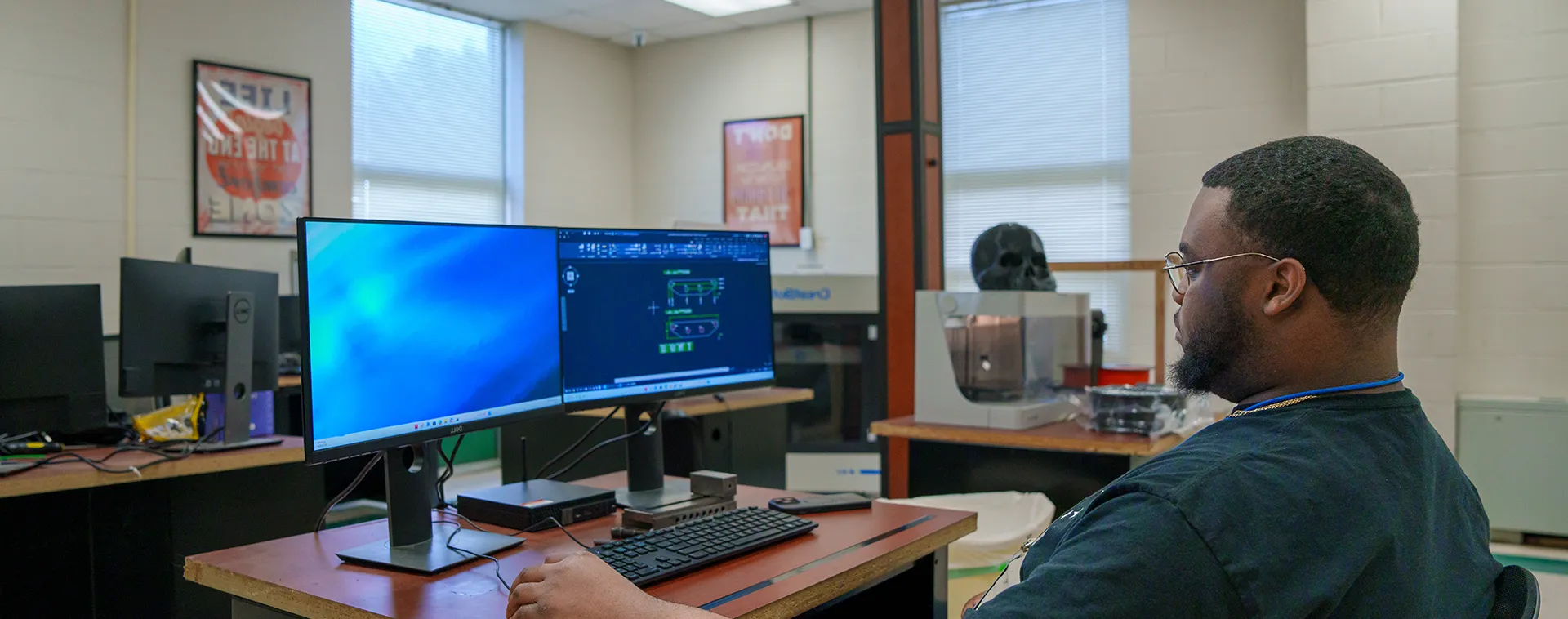 Student seated at a desk using dual computer monitors in a classroom with posters, windows, and equipment behind.