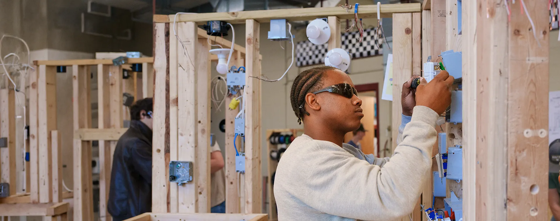 Student working on an electrical training wall, connecting wires to blue electrical boxes mounted on wooden framing.