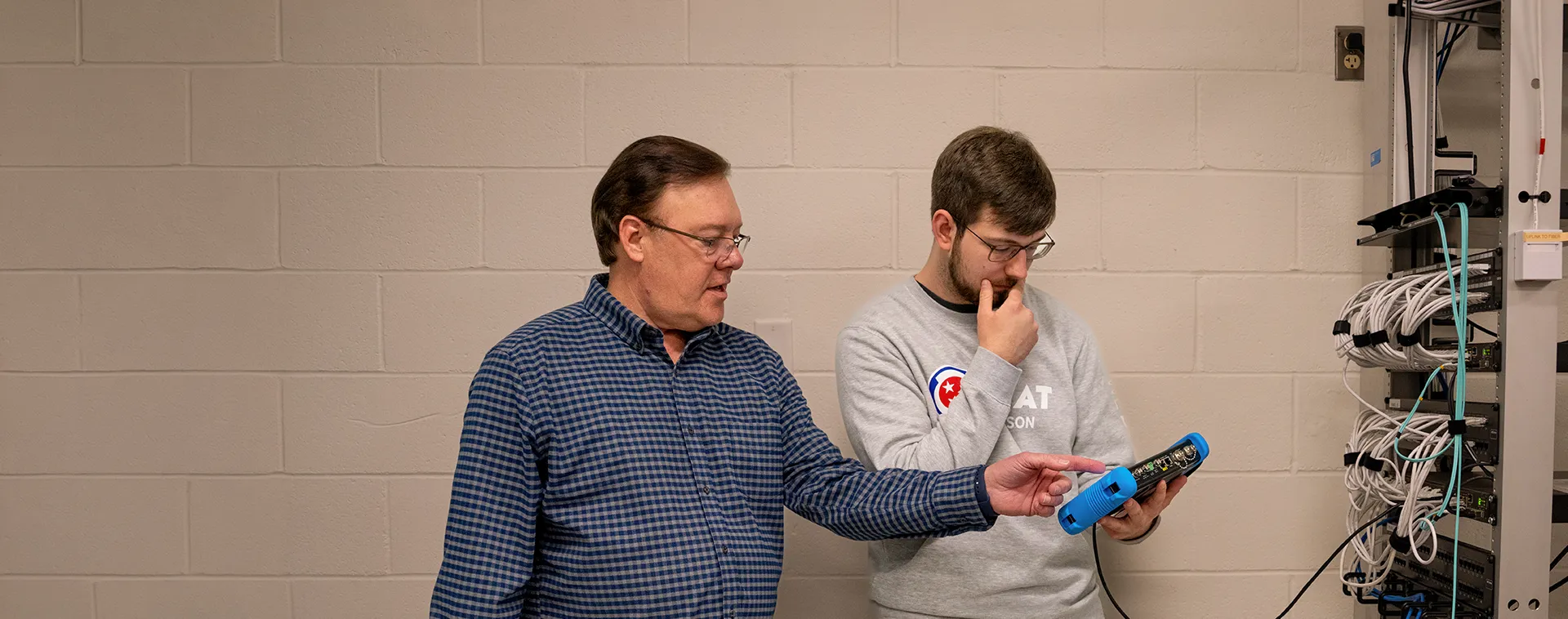 Instructor and student standing beside a network equipment rack, reviewing a handheld testing device and connected cables.