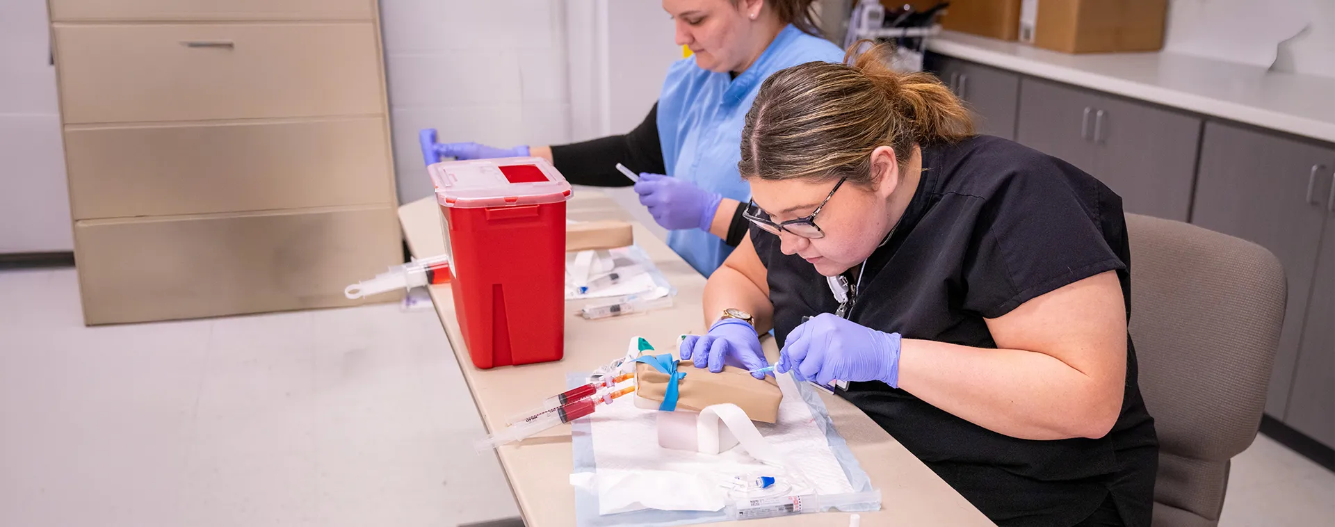 Two students wearing gloves practice injections on a medical training arm at a classroom table with syringes and a sharps container.