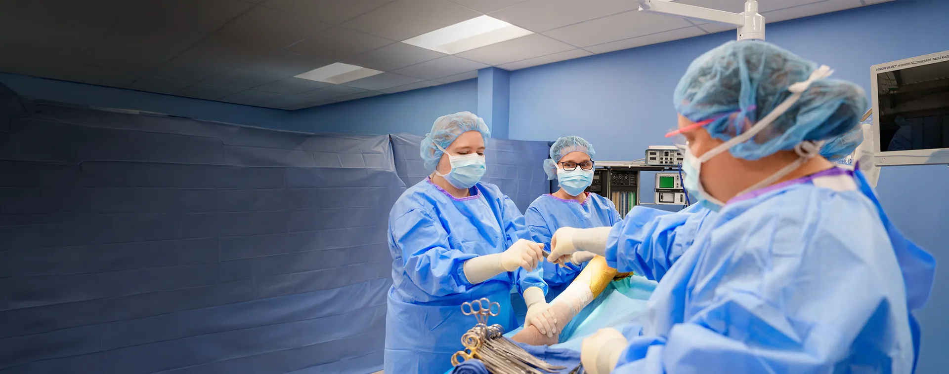 Three people wearing surgical gowns, gloves, and caps stand around a draped operating table in a clinical training room.