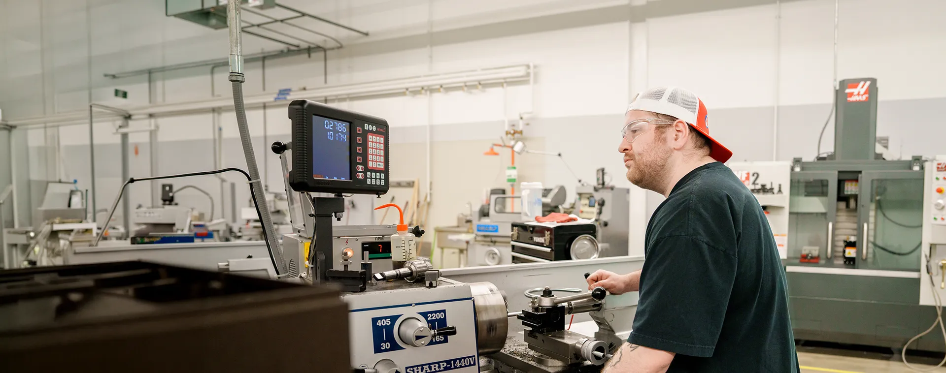 Person operates a metal lathe in a machining lab, adjusting controls with digital readout and industrial equipment nearby.