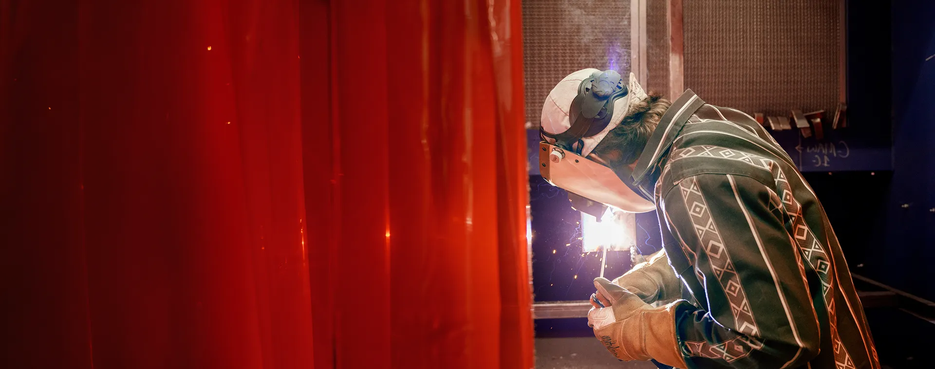 Person wearing a welding helmet and gloves welds metal behind a red safety curtain, with sparks lighting the workshop.
