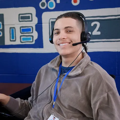 Student wearing a headset seated at a computer desk, with a blue wall mural and cables visible behind.