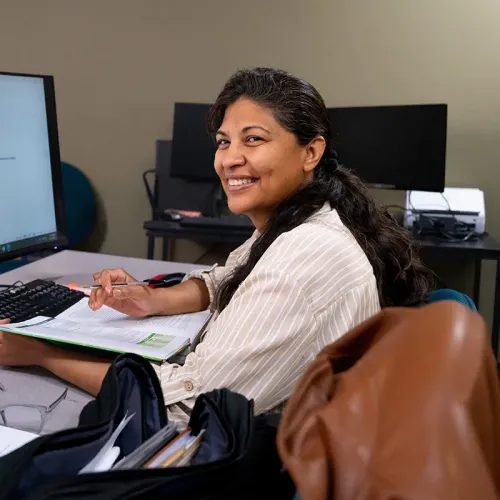 Student seated at a desk reviewing papers beside a computer monitor, with additional workstations in the background.