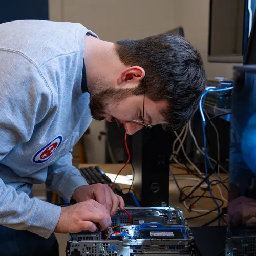 Student working inside an open desktop computer, connecting internal cables at a workstation with monitors nearby.