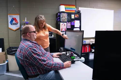 Instructor pointing at a computer monitor while a student sits at a desk in a classroom with cabinets and a whiteboard.