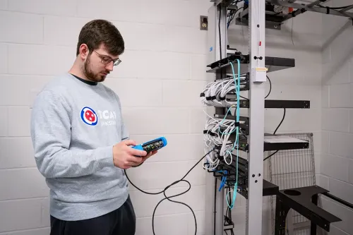 Student holding a network testing device while standing next to a server rack with bundled cables and mounted hardware.