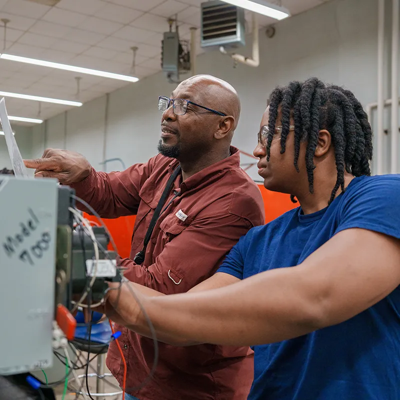 Two students working together on an electrical control panel in a lab with wiring, tools, and equipment.