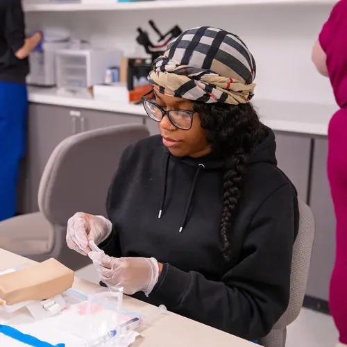 Student wearing gloves prepares medical supplies at a classroom table with training models and cabinets in the background.