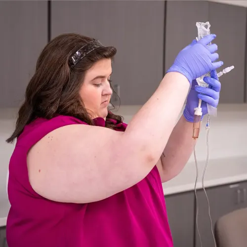Person wearing gloves holds an IV bag and tubing at eye level inside a clinical training room.