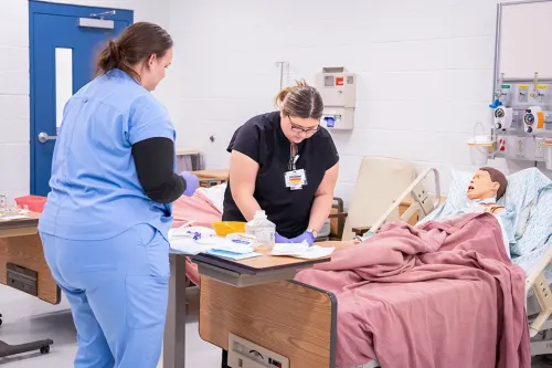Two people stand beside a hospital bed, organizing supplies while practicing patient care in a skills lab.