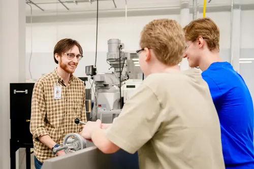 Three people standing at a metalworking machine in a workshop, one demonstrating controls while two observe.
